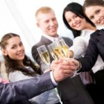 Five people in business attire toasting with champagne glasses, smiling and standing close together in an office setting.