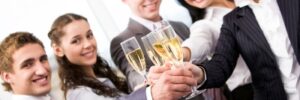 Five people in business attire toasting with champagne glasses, smiling and standing close together in an office setting.