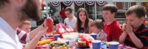 A group of people, including children, sitting at a picnic table outside enjoying a meal with sandwiches and watermelon.