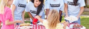 Volunteers at a bake sale table give food to a child. The table is covered with a red and white checkered cloth.