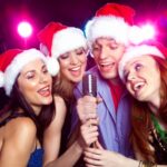 Four people wearing Santa hats sing into a microphone with colorful lights in the background.