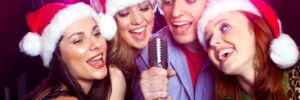 Four people wearing Santa hats sing into a microphone with colorful lights in the background.