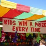 Carnival game booth with a sign reading "Kids Win a Prize Every Time," featuring colorful stuffed animals as prizes.