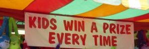 Carnival game booth with a sign reading "Kids Win a Prize Every Time," featuring colorful stuffed animals as prizes.