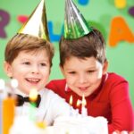 Two children wearing party hats smile at a birthday cake with lit candles in a colorful room.