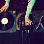 DJ using turntables and audio mixer, adjusting knobs with both hands in a dimly lit setting.