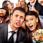 A bride and groom make funny faces with friends posing playfully around them in a group wedding photo.