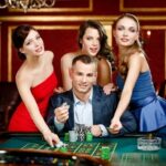 A man in a suit and three women in dresses pose at a casino table with poker chips and cards, set against a lavish interior.