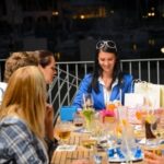 A group of people sitting around a table, enjoying drinks and chatting, with gift bags and glasses on the table.