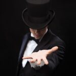 A person in formal attire, wearing a top hat, extends one hand forward against a dark background.