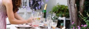 A woman sets a table outdoors with floral decorations, glasses, and a champagne bottle in an ice bucket on a sunny day.