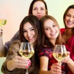 Four women smiling and holding glasses of white wine against a colorful background.