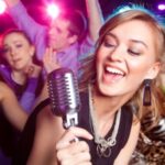 A woman sings into a microphone at a party, surrounded by four people dancing under colorful lights.