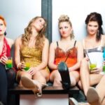 Six women in colorful dresses sit on a couch, holding drinks. They appear relaxed with varied facial expressions.