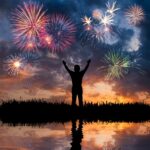 Silhouette of a person with raised arms watching colorful fireworks in the sky above a reflective water surface at twilight.