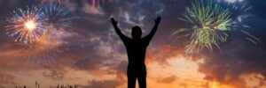 Silhouette of a person with raised arms watching colorful fireworks in the sky above a reflective water surface at twilight.