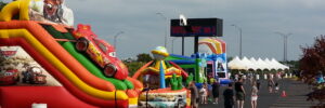 Inflatable slides and attractions set up in a parking lot with people walking around and tents in the background.