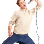 A person kneels while singing passionately into a microphone against a white background.