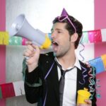 Man in a party hat and suit holding a megaphone and a cup, surrounded by colorful streamers and decorations.