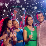 A group of people at a party take a selfie while holding champagne glasses, with festive lights and snow effects in the background.