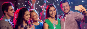 A group of people at a party take a selfie while holding champagne glasses, with festive lights and snow effects in the background.