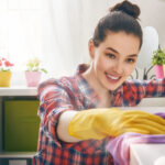 Woman wearing yellow gloves cleans a surface with a purple cloth in a bright room with potted plants.