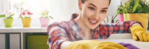 Woman wearing yellow gloves cleans a surface with a purple cloth in a bright room with potted plants.