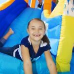 Child smiling while sliding down a blue inflatable slide, with another child climbing behind on a sunny day.