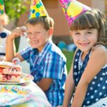 Children at an outdoor party wearing colorful hats, sitting at a table with cake and drinks, and smiling at the camera.