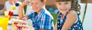 Children at an outdoor party wearing colorful hats, sitting at a table with cake and drinks, and smiling at the camera.