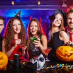 Five people in Halloween costumes smiling at a party with drinks in hand, surrounded by carved pumpkins and decorations.