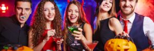 Five people in Halloween costumes smiling at a party with drinks in hand, surrounded by carved pumpkins and decorations.