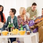 Five people enjoying a casual gathering with balloons, drinks, and snacks; one person is playing a guitar in the living room.