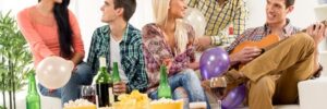 Five people enjoying a casual gathering with balloons, drinks, and snacks; one person is playing a guitar in the living room.