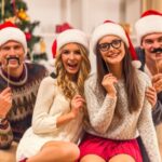 Four people wearing Santa hats, holding fake mustaches, pose in front of a decorated Christmas tree.