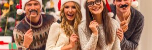 Four people wearing Santa hats, holding fake mustaches, pose in front of a decorated Christmas tree.