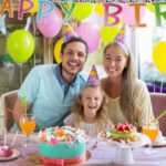 A family of three wearing party hats sits at a table with a birthday cake, fruit tart, and colorful drinks.