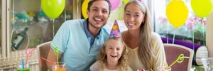 A family of three wearing party hats sits at a table with a birthday cake, fruit tart, and colorful drinks.