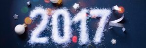 The image shows "2017" written in snow-like material surrounded by baubles, stars, ribbon, and a cork against a dark background.