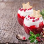 Two dessert glasses with layered red and white parfaits, topped with star-shaped cookies and berries, on a wooden surface.