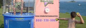 Child aiming ball at man in dunk tank on grassy area by the sea.