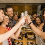 A group of people toasting with glasses while standing around a dining table with plates and a bottle of wine.