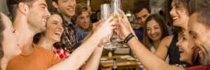 A group of people toasting with glasses while standing around a dining table with plates and a bottle of wine.