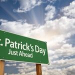 A green sign reading "St. Patrick’s Day Just Ahead" stands against a cloudy sky with sun rays peeking through.