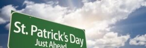 A green sign reading "St. Patrick’s Day Just Ahead" stands against a cloudy sky with sun rays peeking through.