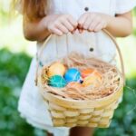 Child holding a wicker basket with colorful eggs and straw, standing on grass.