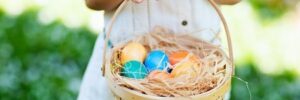 Child holding a wicker basket with colorful eggs and straw, standing on grass.