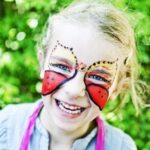 Child with butterfly face paint smiles outdoors, surrounded by greenery.