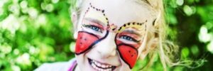 Child with butterfly face paint smiles outdoors, surrounded by greenery.