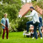 Three men play with a soccer ball on a grassy lawn, with a house and trees in the background.